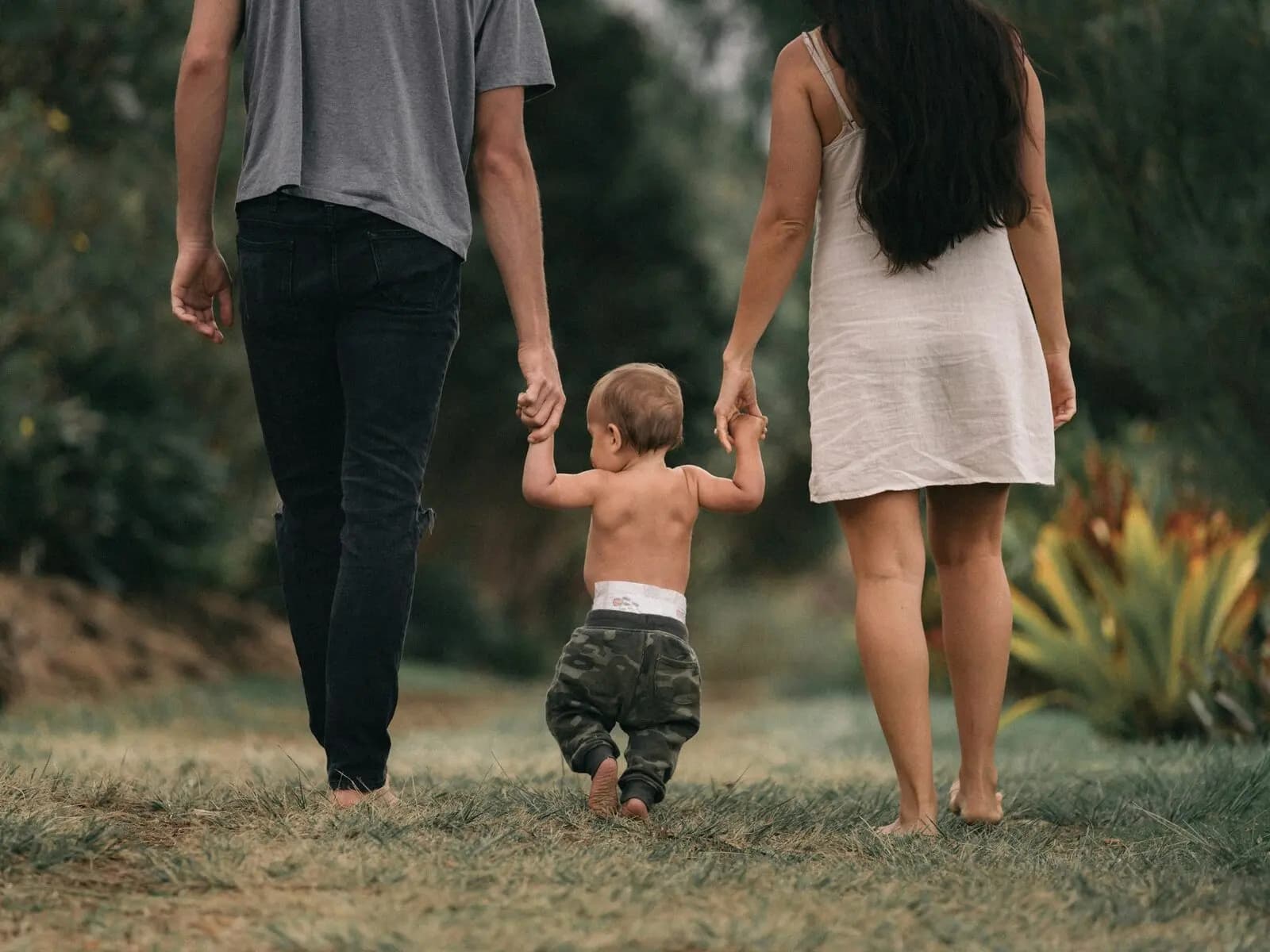 Two parents walking barefoot through a garden, holding their small child by both hands